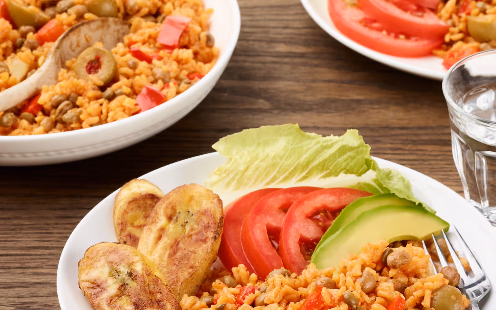 Plate of orange rice with pigeon peas and olives, with fried plantains, avocado, and tomato