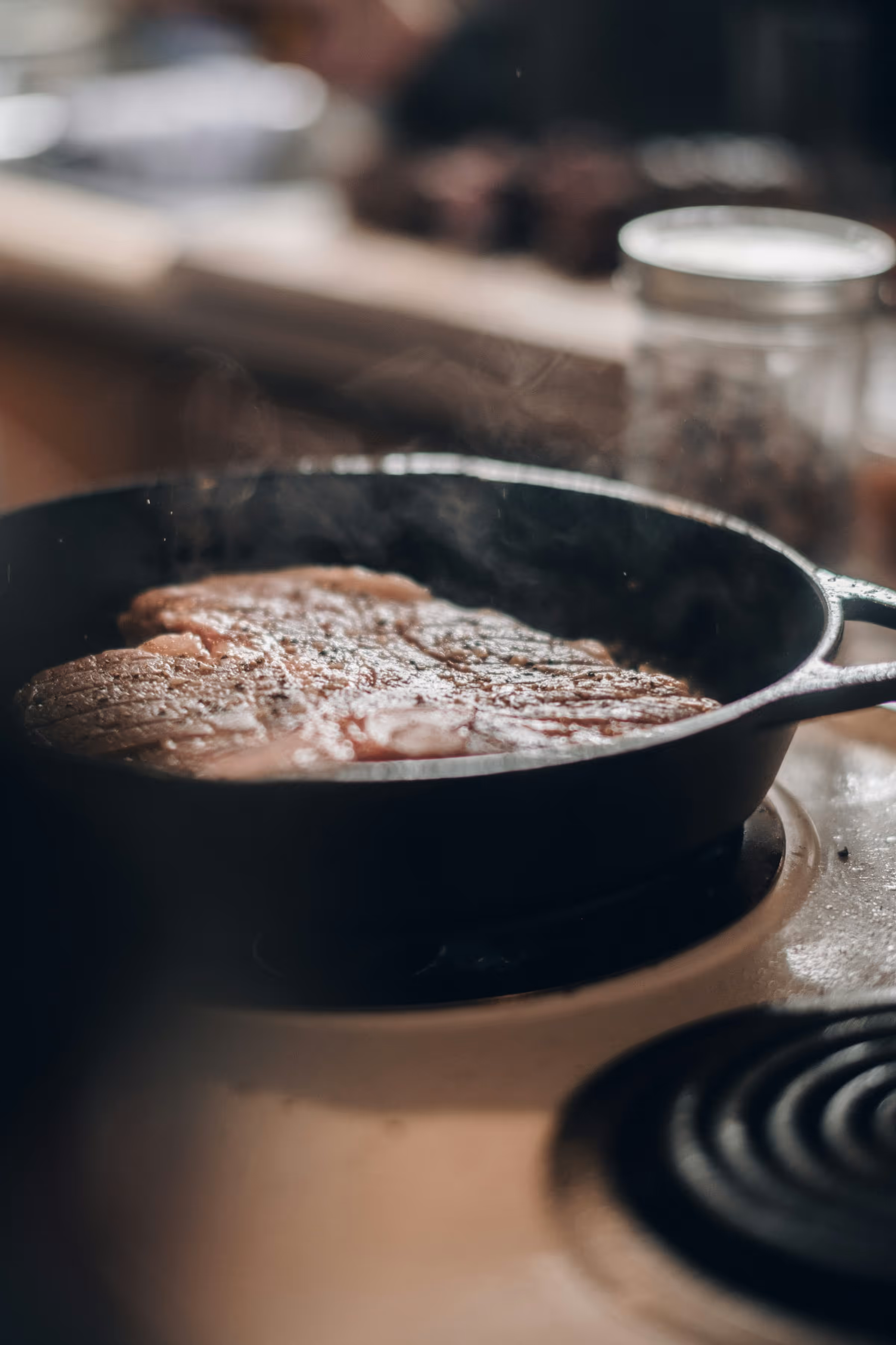Thick steak searing in a black cast-iron skillet on an electric coil, steam rising