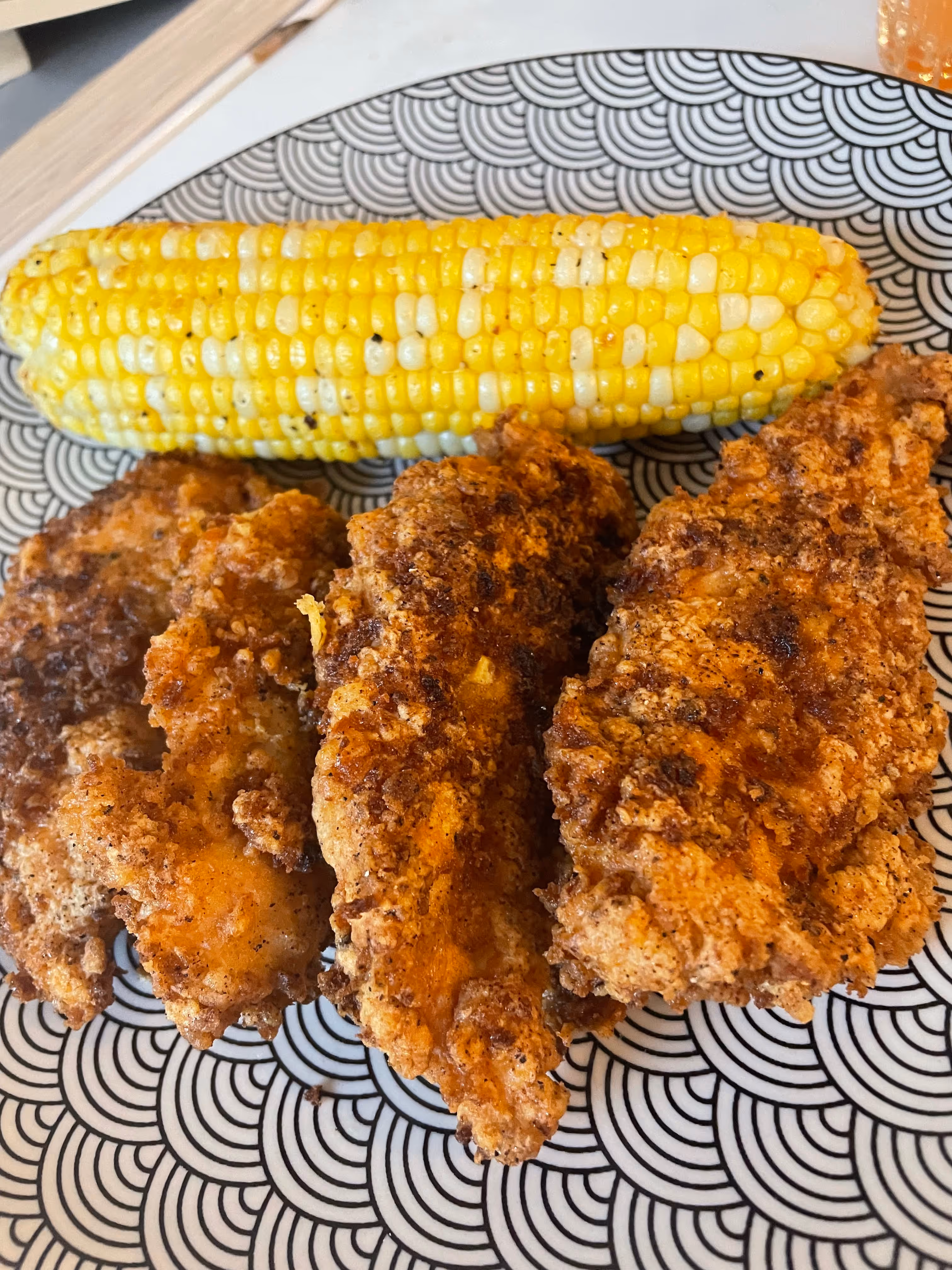 Three fried chicken tenders and a cob of corn on a black-and-white patterned plate