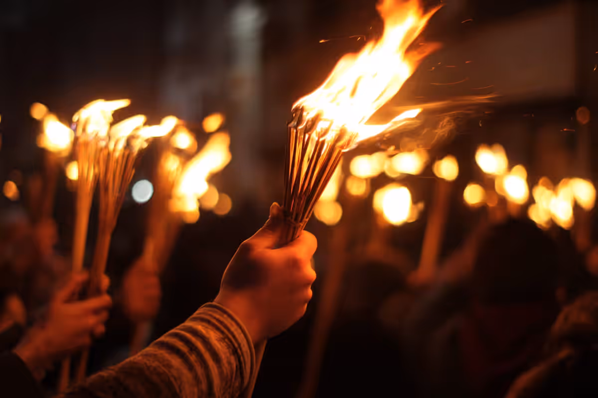 Hand gripping a blazing wooden torch at night, with other torches glowing out of focus in a crowded procession behind it