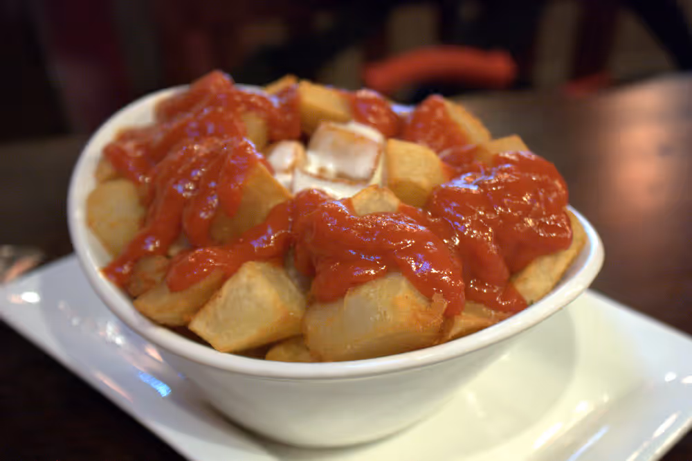 Cubed fried potatoes in a white bowl, red brava sauce and aioli on top
