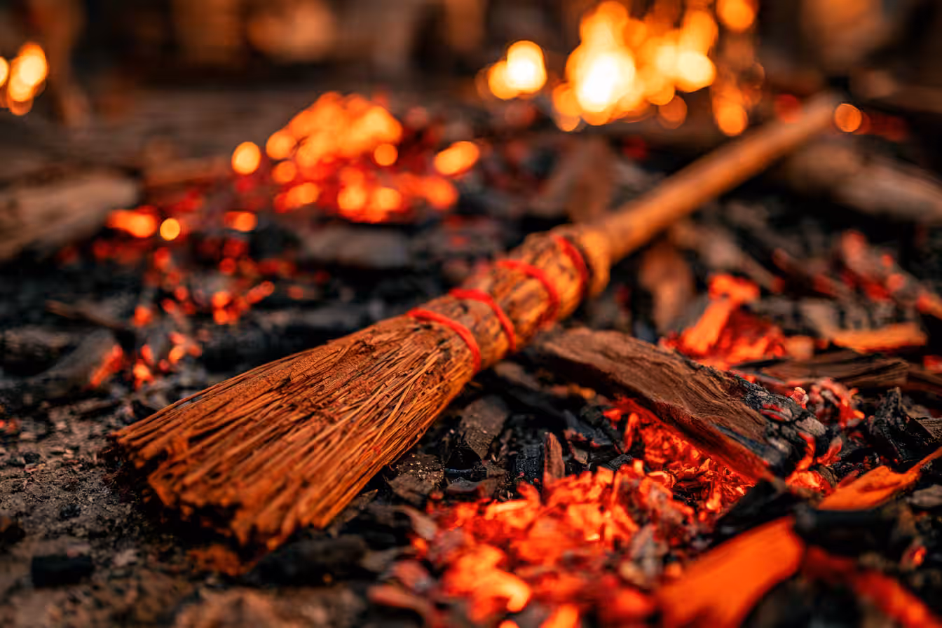 A rustic broom bound with red cord lies across glowing coals and charred wood, with a fire blurred behind it
